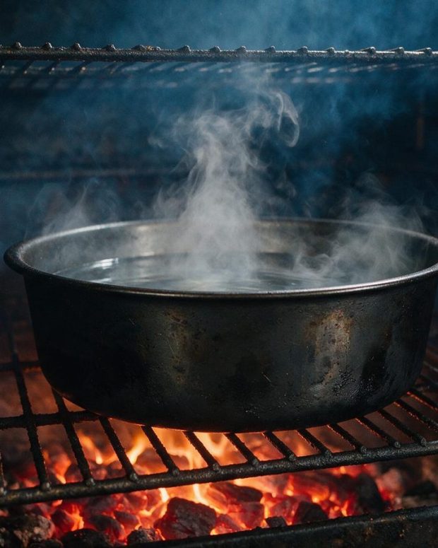 A steel water pan sitting in a smoker chamber, steam rising.